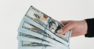 Close-up of a hand holding several stacks of US hundred dollar bills on a white background.